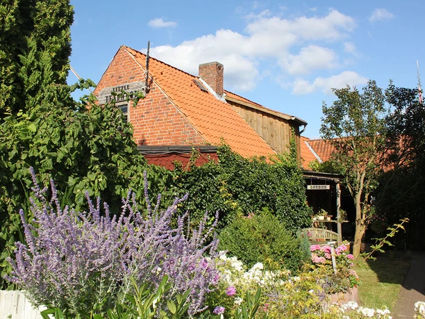 Außenansicht Rotes Backsteinhaus, umgeben von einem Garten mit blühenden Pflanzen und Efeu an der Fassade.