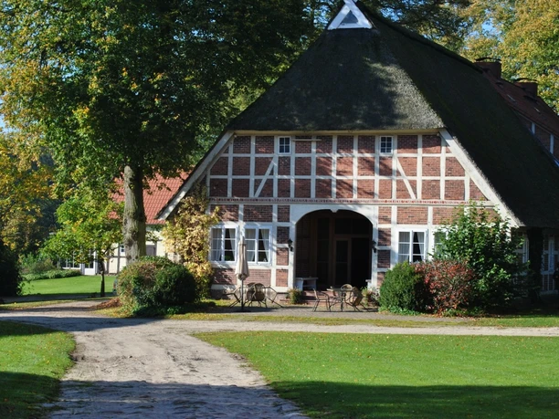 Außenansicht Historisches Fachwerkhaus mit Reetdach auf einem weitläufigen Hofgelände, umgeben von Bäumen und gepflegtem Rasen. Vor dem Gebäude stehen Gartenmöbel auf einer kleinen Terrasse, der Zufahrtsweg ist unbefestigt und führt durch grüne Wiesen bis zum Eingang des Hauses.