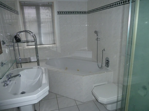 Badezimmer Helles, modern gefliestes Badezimmer mit Eckbadewanne, Dusche mit Glaswand, Waschbecken und WC. Ein Fenster mit Jalousien sorgt für Tageslicht, verchromte Armaturen und Ablagen ergänzen die funktionale Ausstattung.