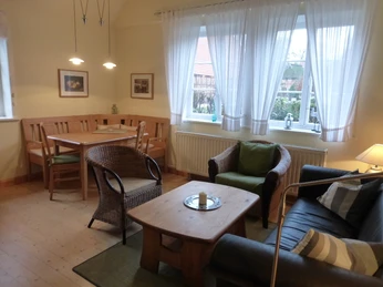 Wohn und Essbereich Heller Wohn- und Essbereich mit großen Fenstern, Holzmöbeln und Sitzbank. Ein schwarzes Ledersofa, zwei Korbstühle und ein Holztisch stehen auf hellem Holzboden. Der Esstisch ist beleuchtet, an den Fenstern hängen weiße Vorhänge mit Blick auf den Garten.