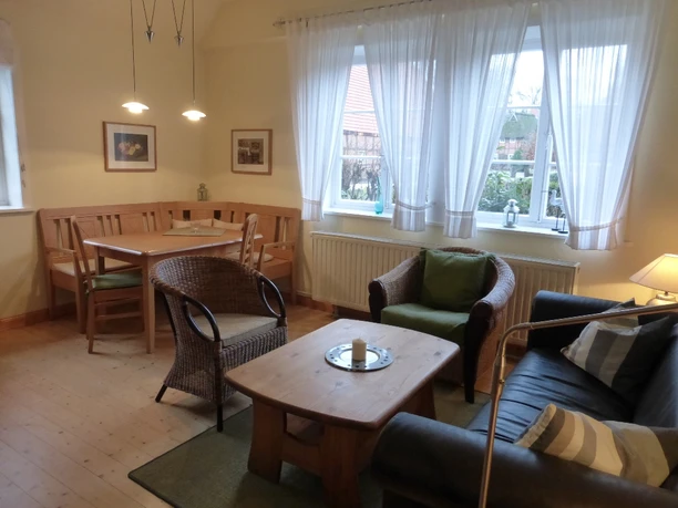 Wohn und Essbereich Heller Wohn- und Essbereich mit großen Fenstern, Holzmöbeln und Sitzbank. Ein schwarzes Ledersofa, zwei Korbstühle und ein Holztisch stehen auf hellem Holzboden. Der Esstisch ist beleuchtet, an den Fenstern hängen weiße Vorhänge mit Blick auf den Garten.