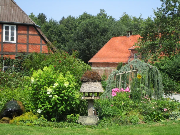 Garten Blühender Bauerngarten mit Reetdach-Vogelhaus und rotem Fachwerkhaus im Hintergrund.