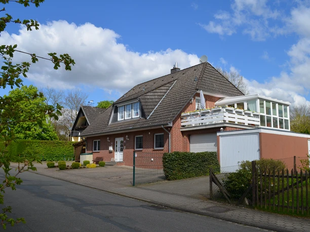 Außenansicht Rotes Backsteinhaus mit dunklem Dach, weißer Haustür, Garage und Balkon im Sonnenlicht.