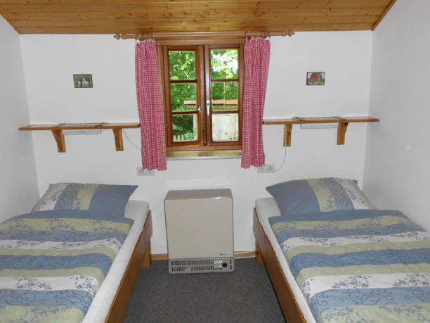 Schlafzimmer Zweibettzimmer mit zwei Holzbetten, gestreifter Bettwäsche in Blau- und Grüntönen, einem Holzfenster mit roten Vorhängen und Blick ins Grüne. Über den Betten befinden sich zwei Wandregale. Der Raum hat eine helle Holzdecke und weiße Wände und vermittelt eine schlichte, funktionale Atmosphäre.