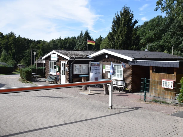 Campingplatz Mühlenkamp Eingangsbereich des Campingplatzes Mühlenkamp mit zwei Holzgebäuden, einem Büro und einer Rezeption, Sitzbänken und Infotafeln. Im Hintergrund stehen Bäume und Wohnwagen auf den Stellplätzen unter blauem Himmel.