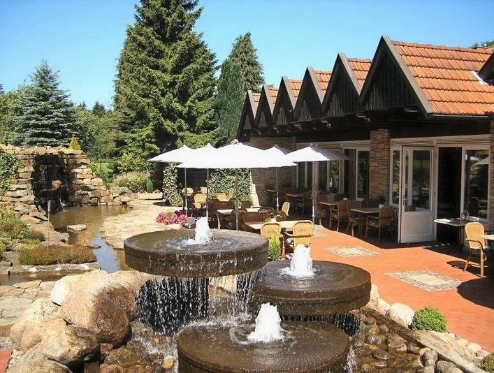 Außenansicht Restauranteingang mit Terrasse, Sonnenschirmen, Springbrunnen und einem kleinen Teich im Grünen.