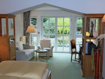 Helles Zimmer mit großen Fenstern, gemütlicher Sitzecke, Arbeitsbereich und Ausblick in den Garten.