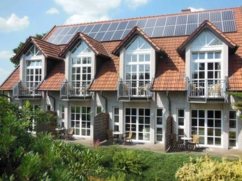 Das Bild zeigt ein zweistöckiges Gebäude mit Ziegeldach und weißen Fensterfronten in grüner Umgebung.