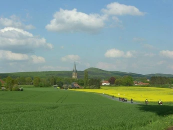 Motorradfahrer fahren an weiten, grünen Feldern vorbei, dahinter ein Dorf mit Kirchturm unter blauem Himmel.