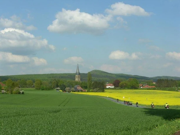 Landschaft Motorradfahrer fahren an weiten, grünen Feldern vorbei, dahinter ein Dorf mit Kirchturm unter blauem Himmel.