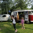 Zwei Kinder spielen auf einem Campingplatz vor einem klassischen roten VW-Bus und einem Wohnwagen.