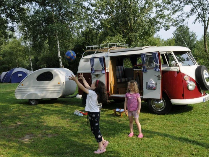 Zwei Kinder spielen auf einem Campingplatz vor einem klassischen roten VW-Bus und einem Wohnwagen.