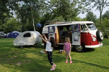 Ferienanlage Regenbogen: Camper Zwei Kinder spielen auf einem Campingplatz vor einem klassischen roten VW-Bus und einem Wohnwagen.