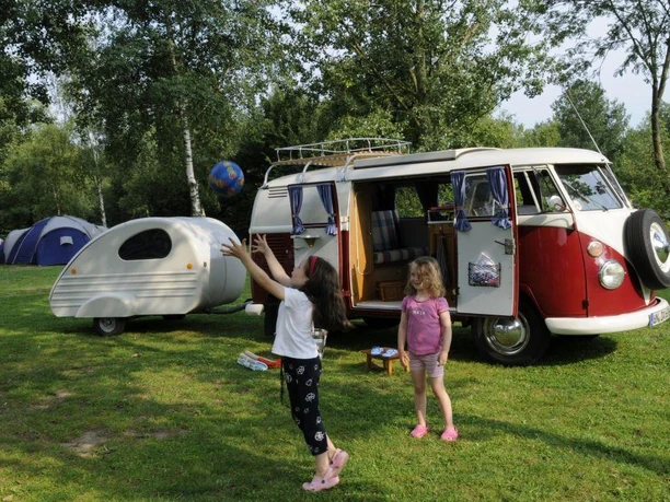 Ferienanlage Regenbogen: Camper Zwei Kinder spielen auf einem Campingplatz vor einem klassischen roten VW-Bus und einem Wohnwagen.