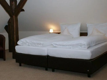 Doppelbettzimmer im Dachgeschoss mit Holzbalken, hellen Wänden, zwei Nachttischlampen und Sessel.