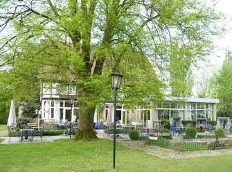 AUßenansicht Saltenhof Hörstel Stilvolles Landhaus mit Terrasse unter einem großen Baum, umgeben von grüner Vegetation und Gartenmöbeln.