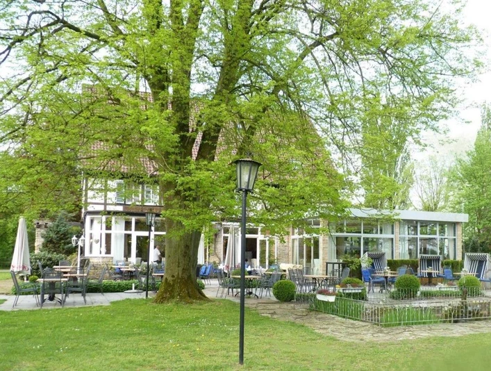 AUßenansicht Saltenhof Hörstel Stilvolles Landhaus mit Terrasse unter einem großen Baum, umgeben von grüner Vegetation und Gartenmöbeln.