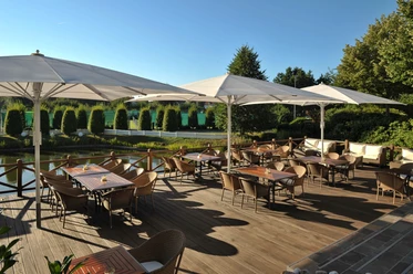 Seeterrasse Restaurantterrasse mit Holzboden, Tischen und Stühlen im Freien, umgeben von grüner Natur.