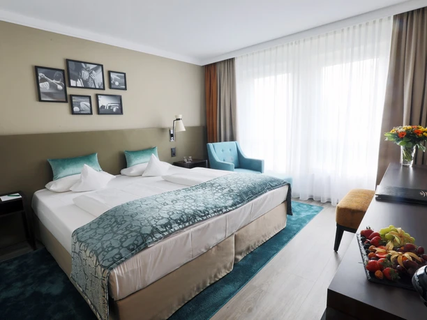 Comfort Deluxe Zimmer im Court Hotel Ein modern eingerichtetes Hotelzimmer mit Doppelbett, blauen Akzenten, Schreibtisch und großen Fenstern.