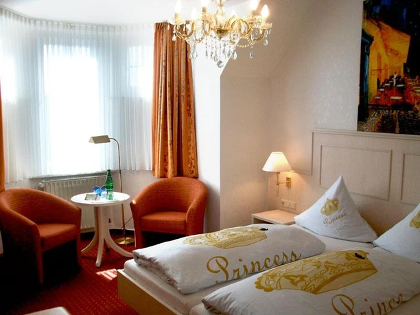 Doppelzimmer Helles Doppelzimmer mit roten Sesseln, Kronleuchter, zwei Betten und einem Gemälde an der Wand.