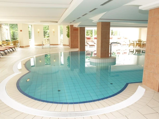 Schwimmbad im Ringhotel Teutoburger Wald in Tecklenburg Brochterbeck Innenansicht eines Schwimmbads mit einem geschwungenen Becken, umgeben von Liegestühlen und großen Fenstern.