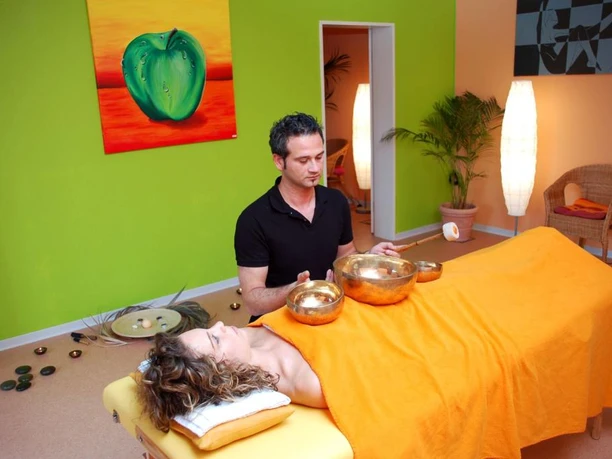 Eine Frau liegt entspannt unter einer orangefarbenen Decke auf einer Liege, während ein Mann mit Klangschalen eine Therapie durchführt.