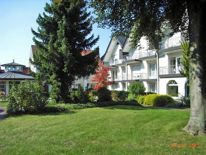 Ferienhotel Becker Ansicht von der Parkseite Weiße Hotelgebäude mit Balkonen, umgeben von gepflegtem Garten und großen Bäumen, unter blauem Himmel.
