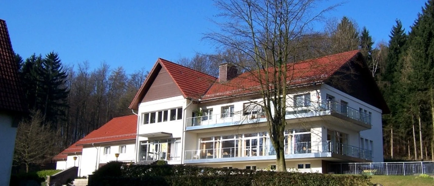 Naturfreundehaus Teutoburg Großes, modernes Gebäude mit rotem Dach, umgeben von Wald und Wiese an einem sonnigen Tag.