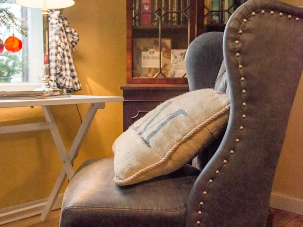 Sessel im Gobelinschlafzimmer Ferienwohnung "Hofgarten" Sessel mit Ziernägeln und Kissen vor Bücherregal, neben Tisch mit Lampe und kariertem Vorhang.