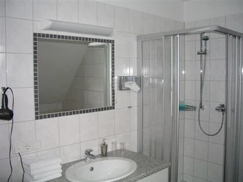 Bad im Hotel Hollmann Modernes Badezimmer mit Dusche, großem Spiegel, Wandföhn und Waschbecken in einem hellen Raum.