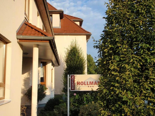 Seitenansicht Hotel Hollmann Hotel Hollmann mit rotem Ziegeldach und Empfangsschild, umgeben von grünen Büschen und Bäumen.