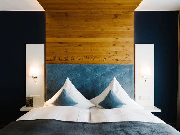 Bett der Hermann Suite im Germanenhof Sandebeck Modernes Hotelbett mit hohen grauen Kopfteil und weißer Bettwäsche, warm beleuchtet.