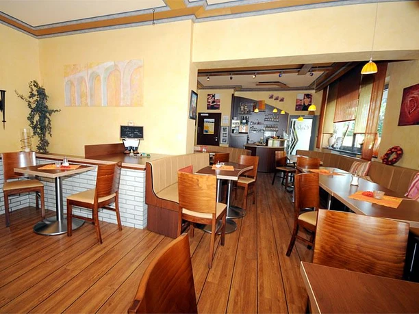 Innenansicht einer hellen Bäckerei mit Holzmöbeln, runden Tischen und großen Fenstern im Hintergrund.