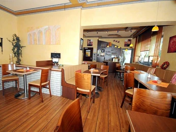 Cafe/Bäckerei Pension Mertens Innenansicht einer hellen Bäckerei mit Holzmöbeln, runden Tischen und großen Fenstern im Hintergrund.