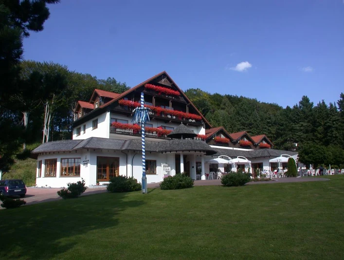 Dreigeschossiges Hotel mit roten Balkongeranien vor bewaldetem Hügel, blauer Himmel im Hintergrund.