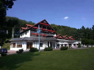 Aussenansicht Hotel Mügge am Iberg***, Oerlinghausen Dreigeschossiges Hotel mit roten Balkongeranien vor bewaldetem Hügel, blauer Himmel im Hintergrund.