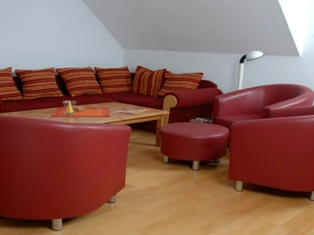 Wohnbereich Roter Wohnbereich mit Sofas und Sesseln, Holzfußboden, Beistelltisch, Lampe und Wandkalender.