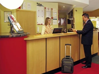 Rezeption Eine Hotelrezeption mit einem Gast, der mit einem Mitarbeiter spricht; ein kleiner Koffer steht daneben.