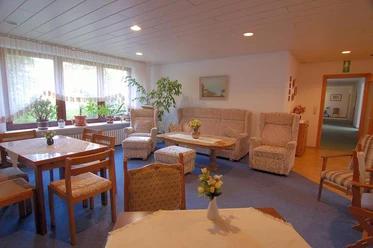 Sitzecke Pension Haus Stork Heller Gemeinschaftsraum mit Sesseln, Sofas, Holztisch und großen Fenstern, durch die Tageslicht fällt.