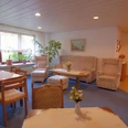 Sitzecke Pension Haus Stork Heller Gemeinschaftsraum mit Sesseln, Sofas, Holztisch und großen Fenstern, durch die Tageslicht fällt.