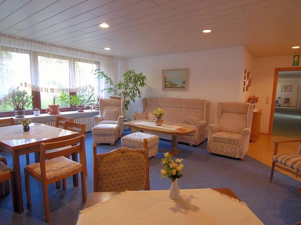 Sitzecke Pension Haus Stork Heller Gemeinschaftsraum mit Sesseln, Sofas, Holztisch und großen Fenstern, durch die Tageslicht fällt.