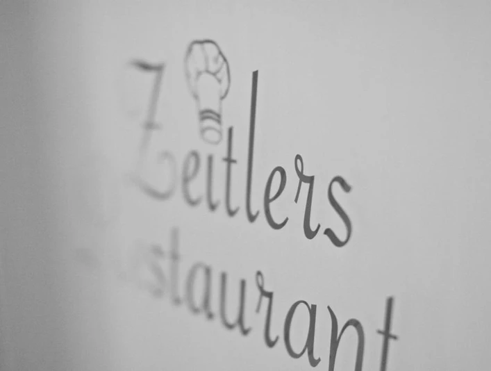 Eingangs-Logo Zeitlers Hotel & Apartments Schwarz-weiß-Fotografie des Schriftzugs "Zeitlers Restaurant" mit einem Kochmützen-Symbol.