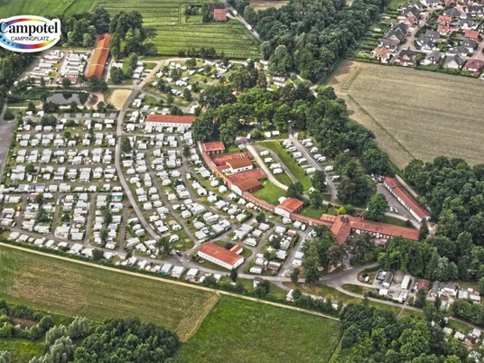 Luftaufnahme eines weitläufigen Campingplatzes mit zahlreichen Wohnwagen und umgeben von Wäldern und Feldern.