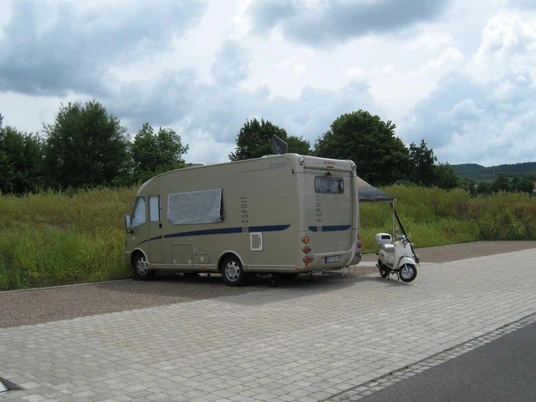 Wohnmobil neben Roller auf gepflastertem Stellplatz, umgeben von Wiesen und Bäumen unter Wolkenhimmel.