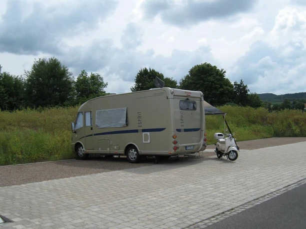 Wohnmobil neben Roller auf gepflastertem Stellplatz, umgeben von Wiesen und Bäumen unter Wolkenhimmel.
