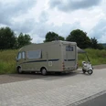 Wohnmobilstellplatz am Bahnhof Bad Holzhausen Wohnmobil neben Roller auf gepflastertem Stellplatz, umgeben von Wiesen und Bäumen unter Wolkenhimmel.