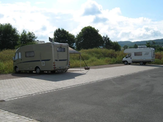 Wohnmobilstellplatz am Bahnhof Bad Holzhausen Zwei Wohnmobile stehen auf einem gepflasterten Stellplatz bei Büschen und Bäumen unter wolkigem Himmel.