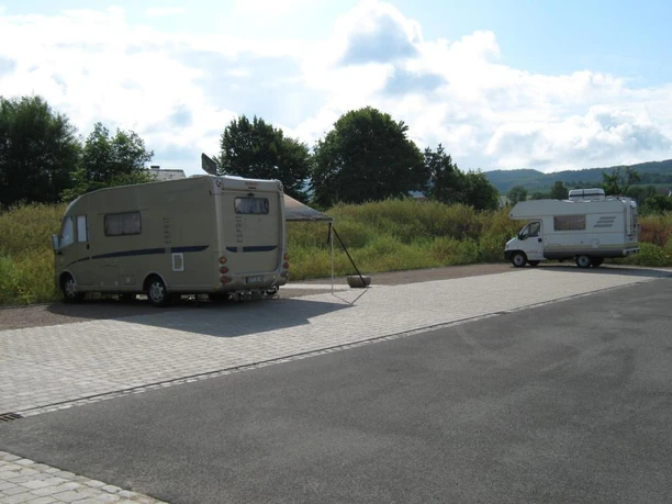 Wohnmobilstellplatz am Bahnhof Bad Holzhausen Zwei Wohnmobile stehen auf einem gepflasterten Stellplatz bei Büschen und Bäumen unter wolkigem Himmel.