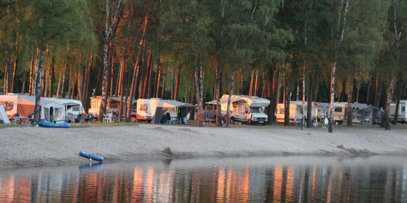 Erholungsanlage Hertha-See Hörstel Wohnwagen und Zelte am sonnigen Ufer eines Sees, umgeben von hohen, dichten Kiefern.