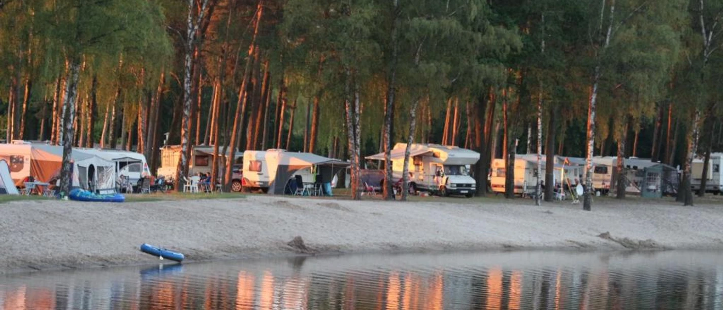 Erholungsanlage Hertha-See Hörstel Wohnwagen und Zelte am sonnigen Ufer eines Sees, umgeben von hohen, dichten Kiefern.